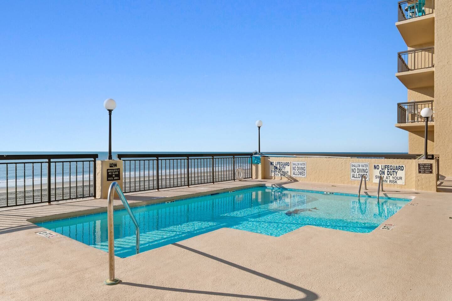 Una piscina con vista a la playa, rodeada por una valla y varios letreros de advertencia.