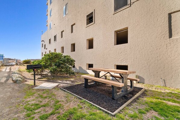 A building with a flat facade, multiple windows, and a picnic table in the foreground. - Oceanfront 1BR Condo with Corner Balcony, Pool (North Myrtle Beach)