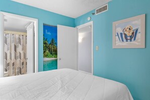 A bedroom with a bed, a door leading to a balcony with a view of palm trees and water, and a framed picture on the wall.
