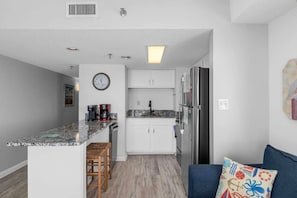 A compact kitchen with white cabinets, a stainless steel refrigerator, a coffee maker, and a clock on the wall. - Oceanfront 1BR Condo with Corner Balcony, Pool (North Myrtle Beach)