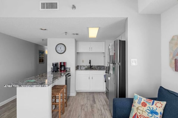 Una cocina compacta con armarios blancos, un refrigerador de acero inoxidable, una cafetera y un reloj en la pared.