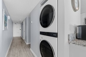 A laundry room with two stacked washing machines, a countertop with a sink, and a hallway leading to another room. - Oceanfront 1BR Condo with Corner Balcony, Pool (North Myrtle Beach)