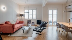 Una sala de estar moderna con un sofá, una silla, una mesa de centro y un televisor. Hay ventanas y una puerta que da a un espacio exterior.