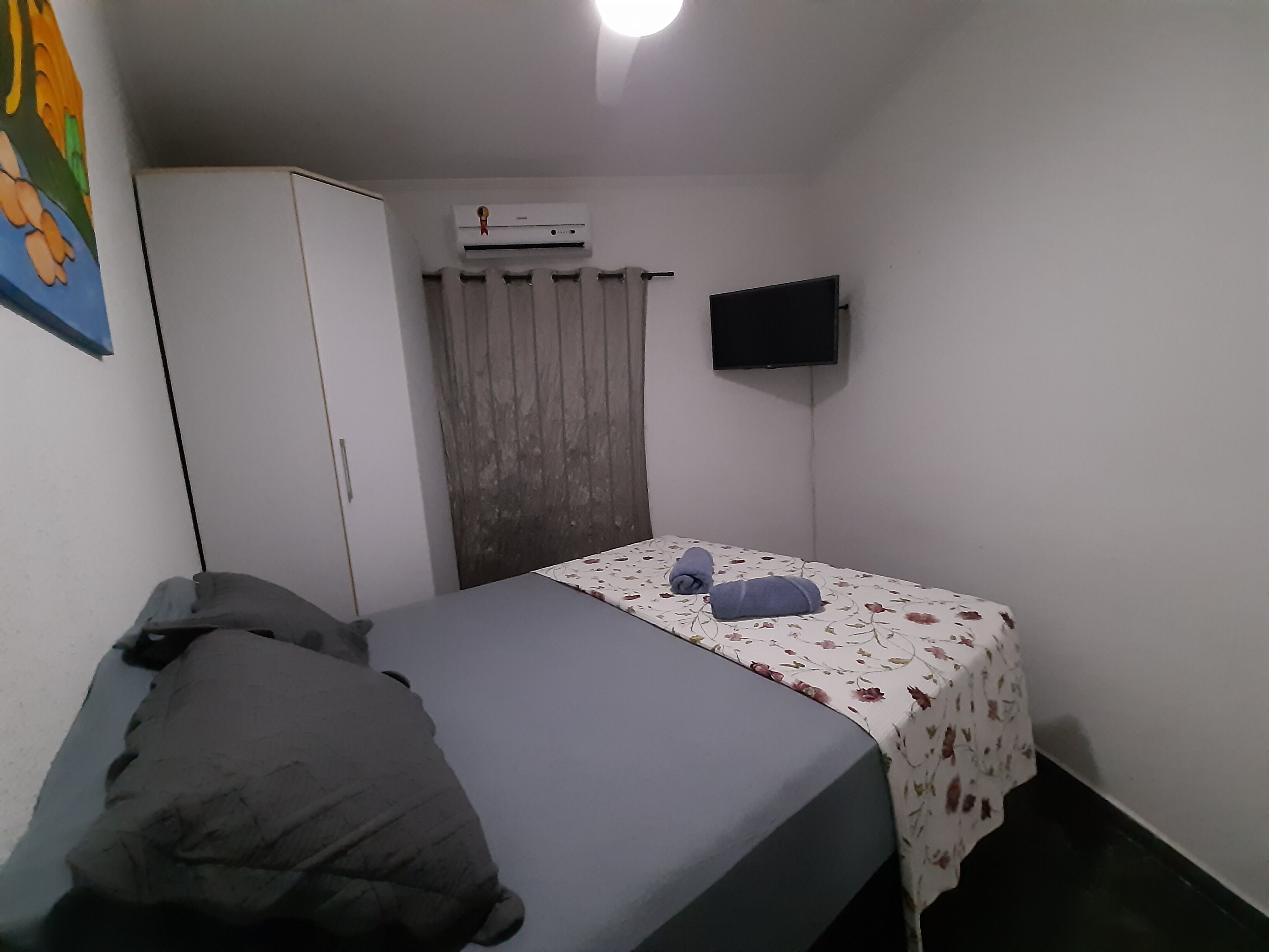Quarto pequeno e simples com cama, guarda-roupa, televisão de tela plana e janela com cortinas.
