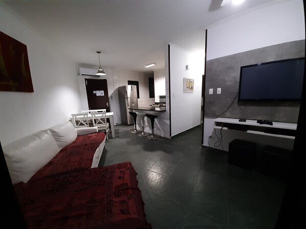 A modern hotel room with a bed, a desk, a chair, a TV, and a kitchenette. - Apartamento com Piscina a uma Quadra da Praia (Vila Clais Bertioga)