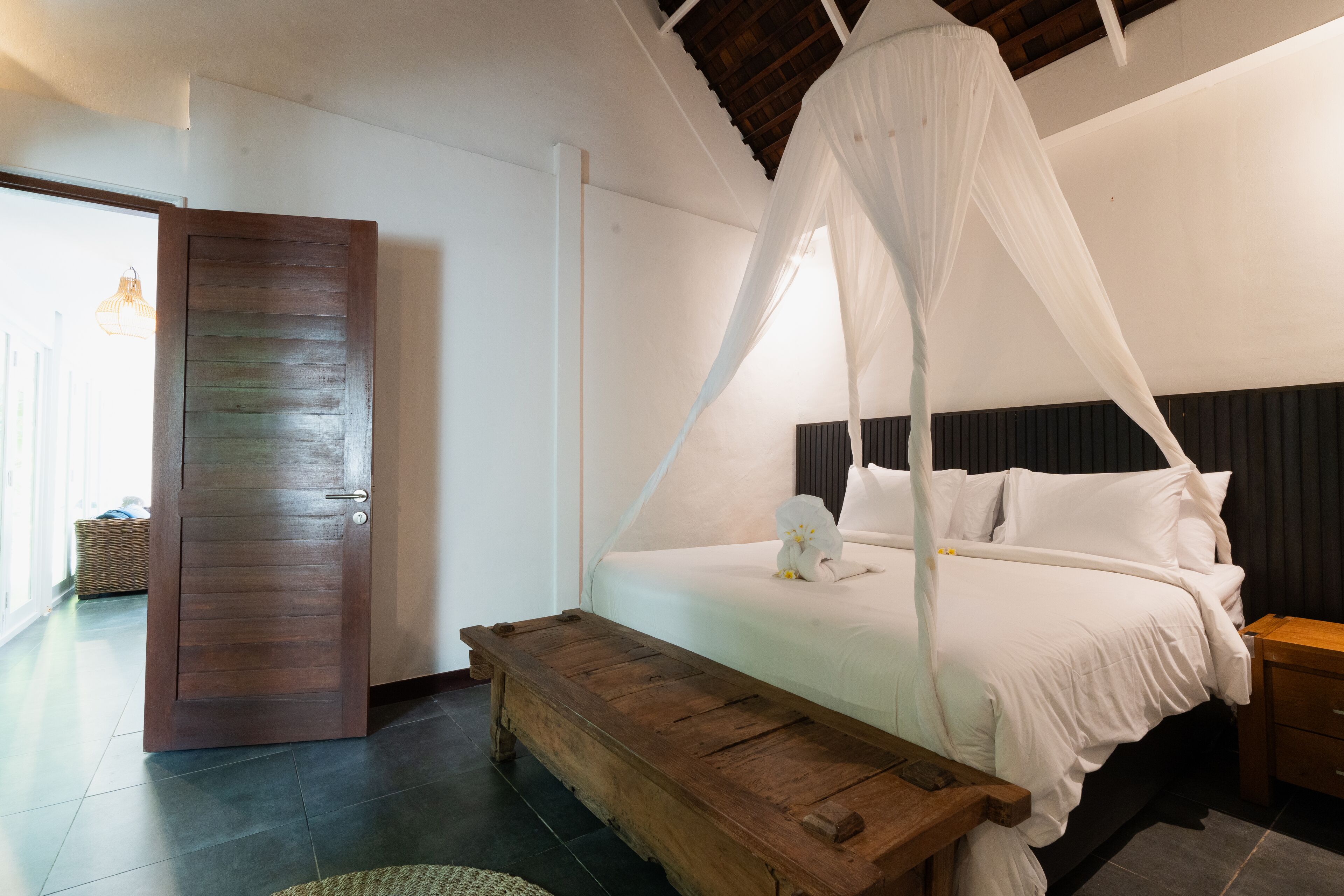 A bedroom with a bed under a canopy, a wooden bench, a nightstand, and a door.
