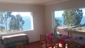 Ein Zimmer mit zwei Betten, einem Tisch und Stühlen. Große Fenster bieten Blick auf das Meer und Bäume.