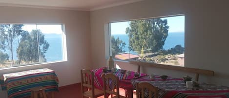 A room with two beds, a table, and chairs. There are large windows with a view of the sea and trees.
