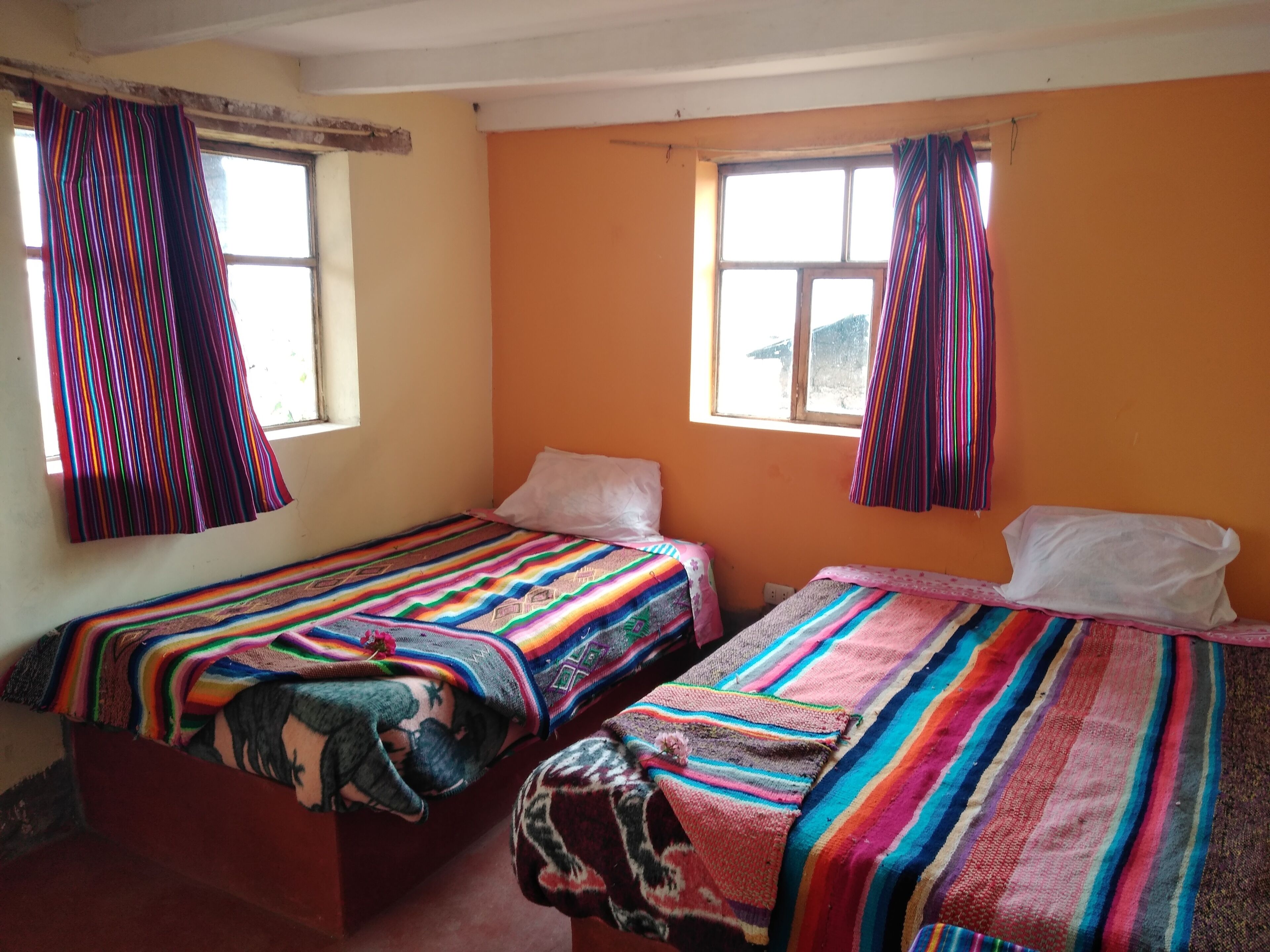 Quarto com duas camas, cada uma com roupa de cama listrada e colorida. Janelas com cortinas, e as paredes pintadas de laranja.