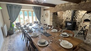 Un coin repas rustique avec une longue table en bois dressée pour le repas, des murs en pierre et une cheminée en pierre.