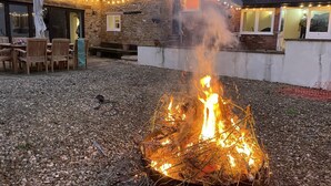 Un bâtiment en pierre avec un foyer au premier plan, des sièges en plein air et des guirlandes lumineuses.
