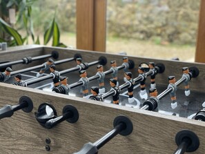 A foosball table with multiple rods and figures, set against a wooden backdrop with a view of greenery outside. - Lovely 5-bed House Near Ilminster and for Families (Chard)