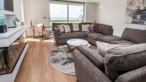 Ein modernes Wohnzimmer mit einem Ecksofa, einem runden Couchtisch und einem großen Fenster mit Blick ins Freie.