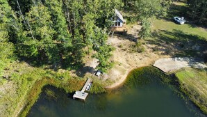 Vista aèria d'una propietat a la vora d'un llac, amb un moll, una cabana i un vehicle aparcat a prop.