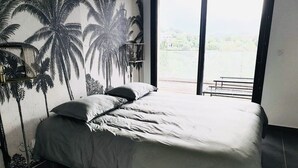 A bedroom with a bed, a nightstand, a large window, and a view of the outdoors. - Appartement 2 Pièces Porto Vecchio (Porto-Vecchio)