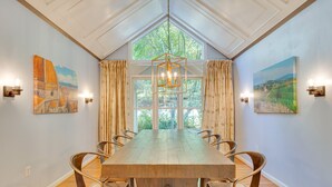 A dining room with a wooden table and chairs, large windows with curtains, and wall-mounted light fixtures. - Blue Ridge Mountain Retreat: Deck & Sunroom! (Fletcher)