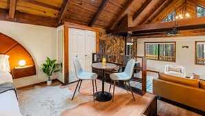 L’intérieur chaleureux d’un chalet avec un plafond en bois, un mur en pierre, un coin repas avec deux chaises, un canapé et une grande fenêtre donnant sur la forêt.