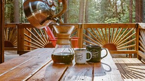 Une terrasse en bois avec une table, une bouilloire en métal, une carafe en verre et deux tasses.