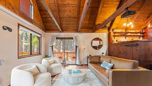 Un salon chaleureux avec un plafond en bois, un canapé, une table basse et une vue sur une terrasse aménagée avec des sièges d’extérieur.