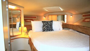 A well-lit, compact bedroom with a large bed, a bedside table with lamps, a mirror, and a small stool.