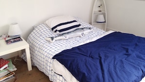 Una cama cuidadosamente tendida con una colcha azul y blanca a cuadros, un almohadón blanco con rayas azules y una mesita de noche blanca con una lámpara y libros.