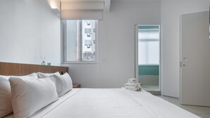 Quarto de hotel minimalista com uma cama grande, uma janela com vista para prédios e uma porta que dá para uma varanda.