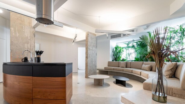 Uma sala de estar moderna com um aparador curvo de madeira, uma mesa de centro redonda, um sofá bege e uma janela grande com vegetação.