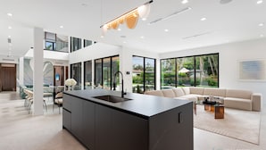 Een moderne keuken met een centraal eiland, grote ramen en een eethoek met een glazen tafel en stoelen.