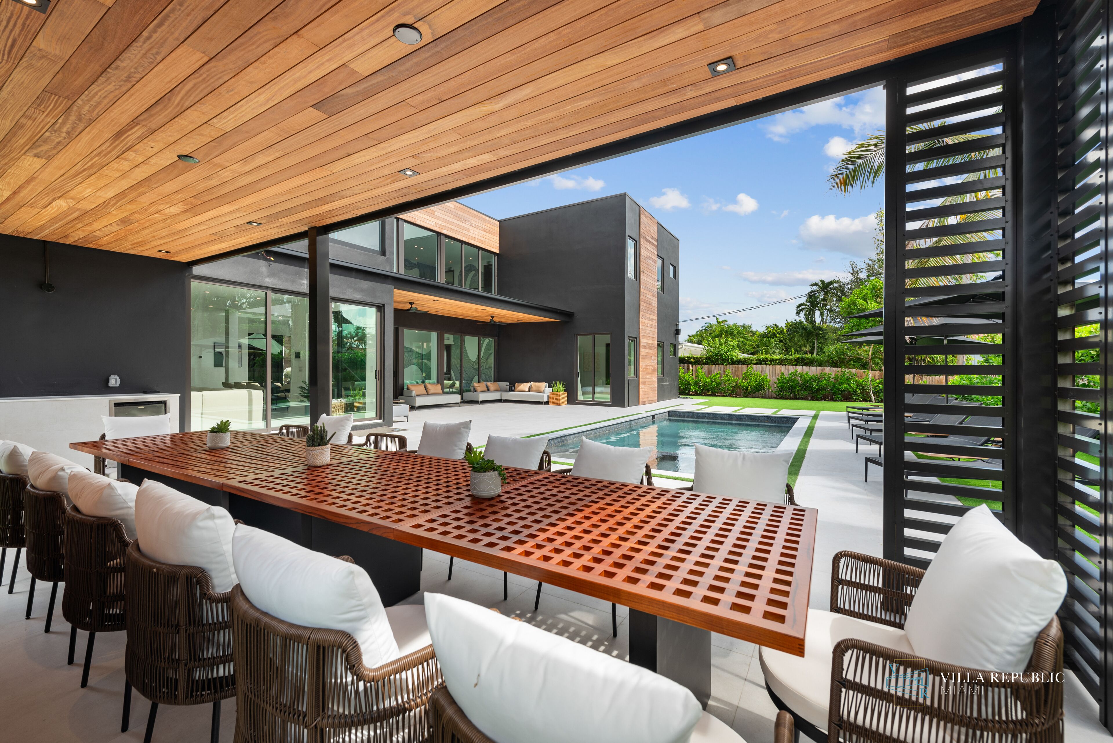 Un espace repas extérieur moderne, avec une table en bois et des chaises en osier, offrant une vue sur une piscine et une végétation luxuriante.