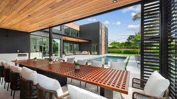Un espace repas extérieur moderne, avec une table en bois et des chaises en osier, offrant une vue sur une piscine et une végétation luxuriante.