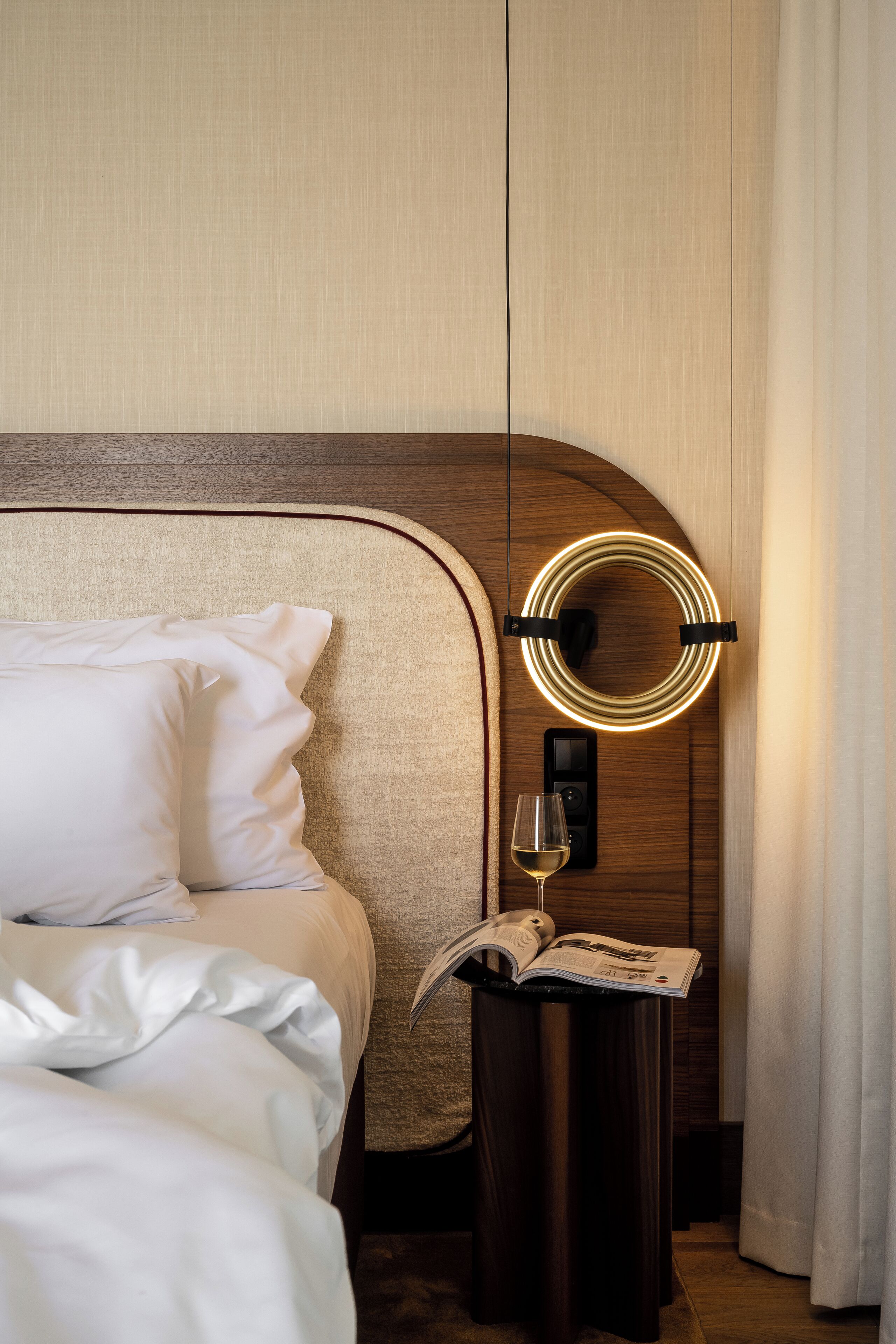A neatly made bed with white linens, a bedside table with a glass of wine and an open magazine, and a modern wall-mounted light fixture.