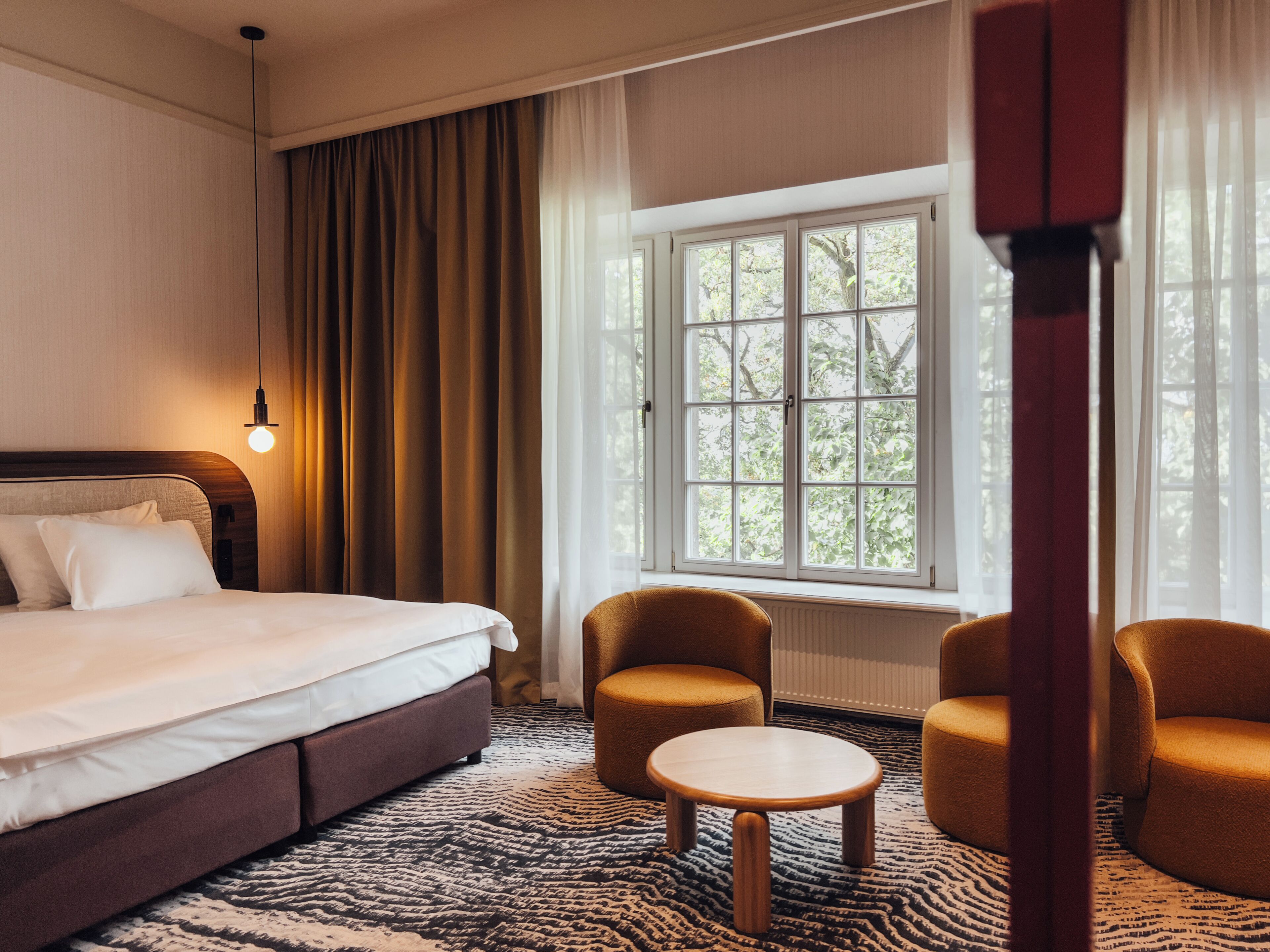 A hotel room with a bed, two armchairs, a small table, a window with a view of trees, and a hanging light fixture.
