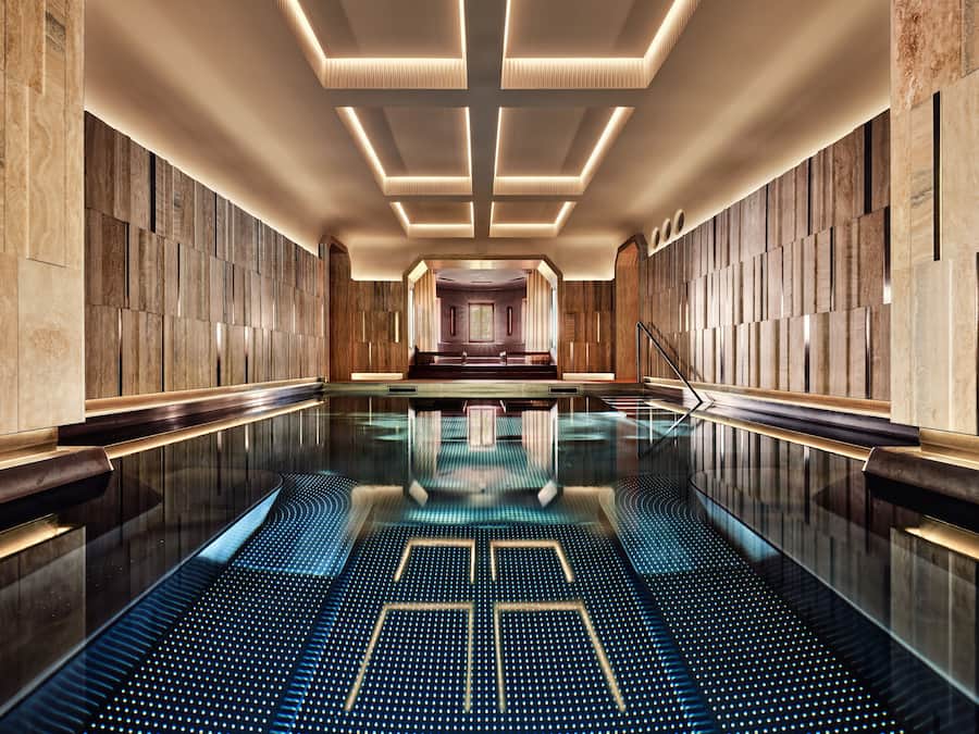 Una piscina interior moderna con un patrón geométrico en el suelo, rodeada de paneles de madera y un área de estar central.