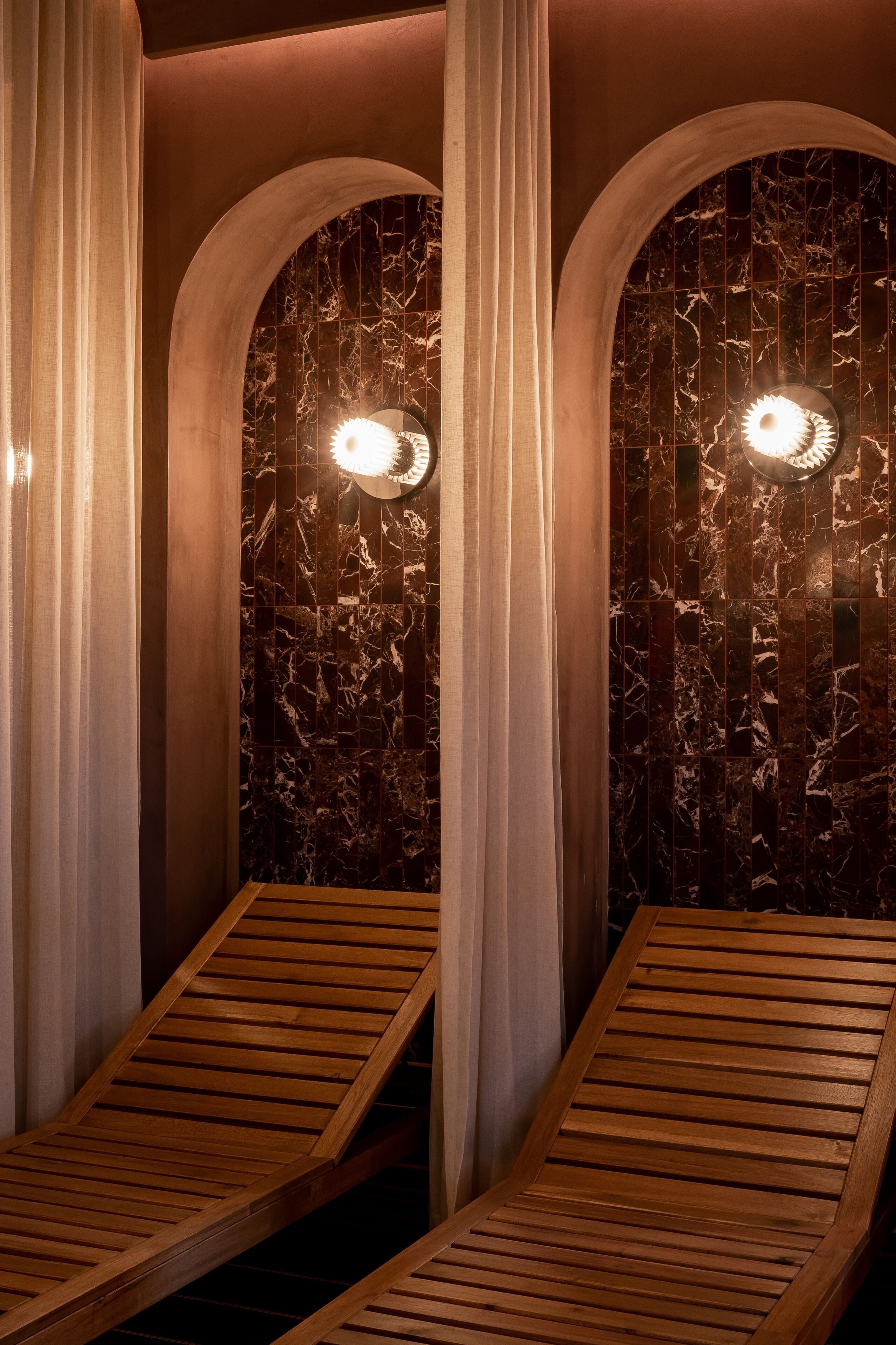 A spa room with two wooden loungers, marble walls, and two wall-mounted lights.