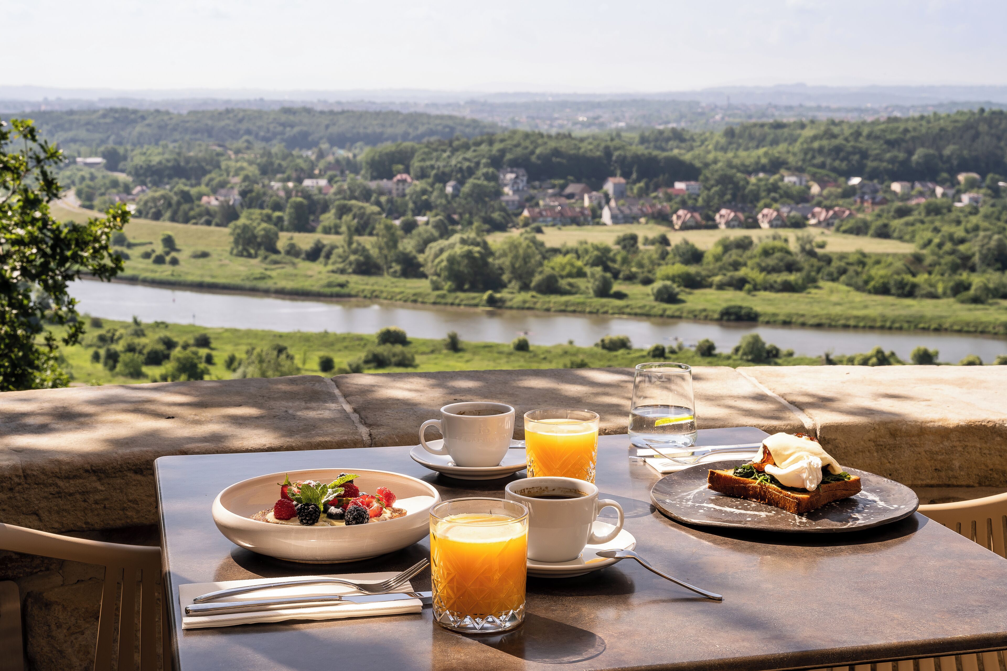Une table dressée avec des aliments pour le petit-déjeuner et des verres de jus d’orange, donnant sur une rivière et un paysage verdoyant.