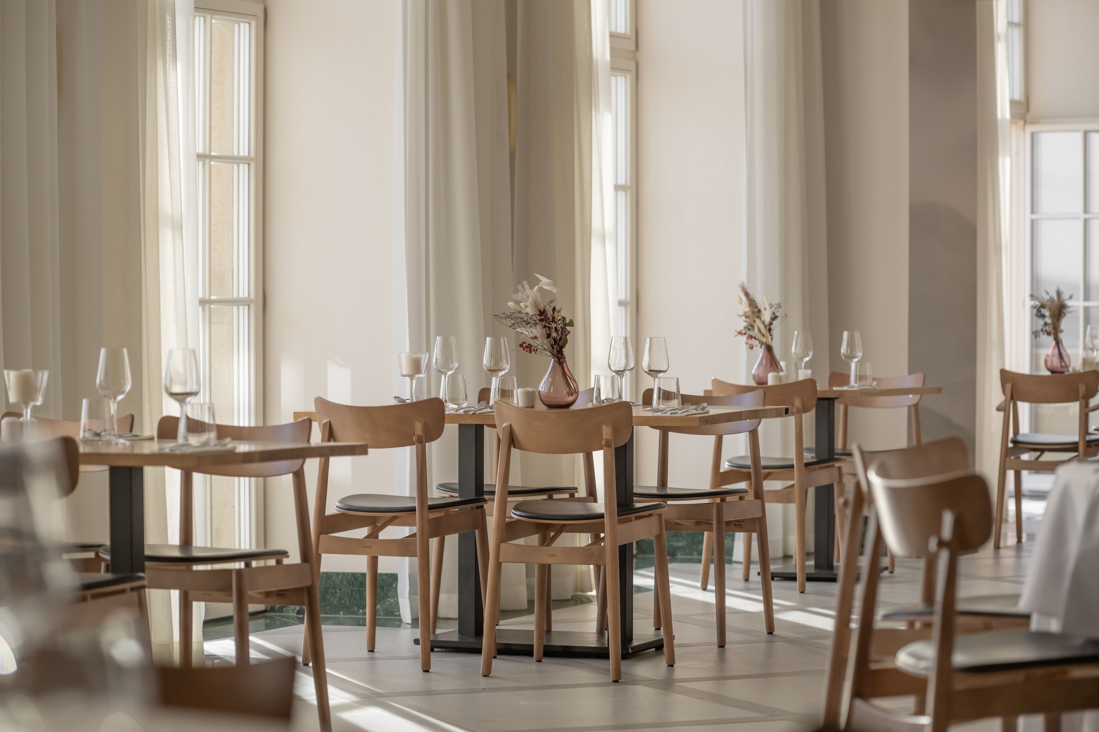 Un comedor con mesas y sillas de madera, dispuesto en una habitación amplia con ventanas grandes que dejan entrar luz natural.