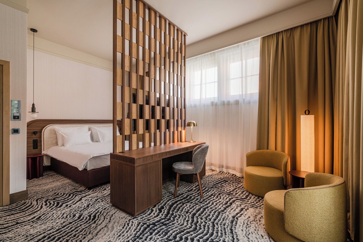 A hotel room with a bed, desk, two chairs, a wooden partition, and a patterned carpet.
