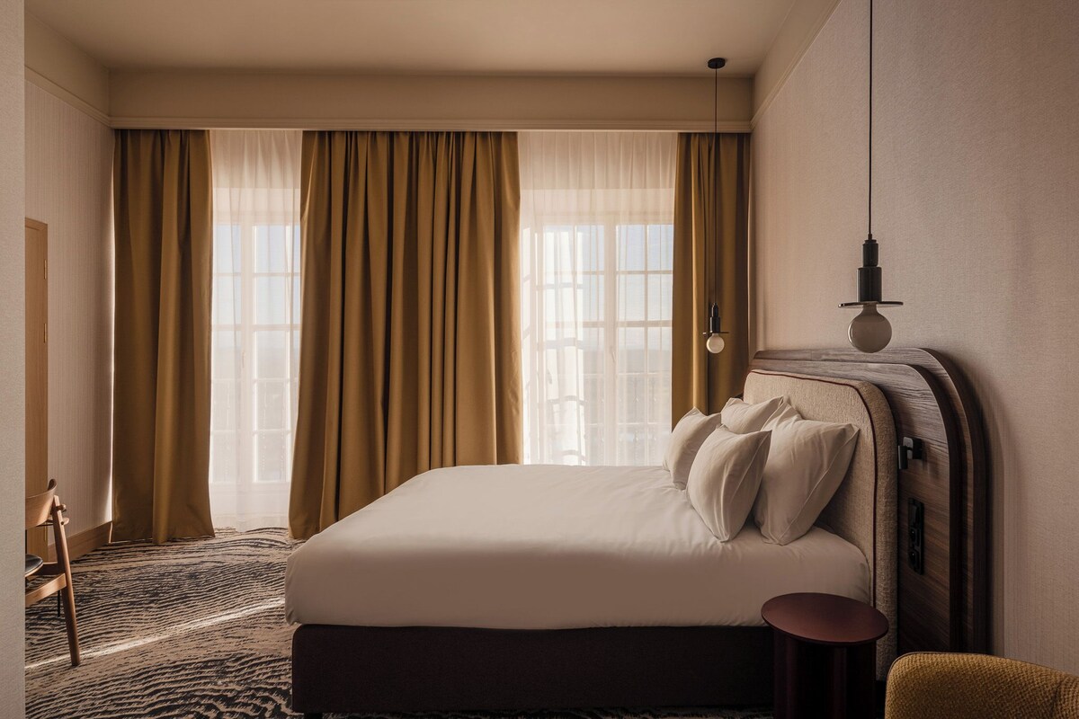 Une chambre d’hôtel avec un grand lit, une chaise, une petite table et une fenêtre avec des rideaux.