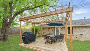 A wooden pergola with a table and chairs underneath, a barbecue grill, and a brick wall in the background. - Game-Day Getaway NEW Stadium View Walk to ATT or Rangers (Arlington)