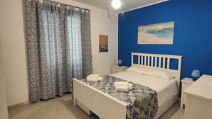 A bedroom with a bed, a nightstand, a desk, a chair, a window with curtains, and a wall painting. - Dolceluna Apartment - San Vito Lo Capo (TP) (San Vito Lo Capo)