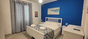 A bedroom with a bed, a nightstand, a desk, a chair, a window with curtains, and a wall painting.