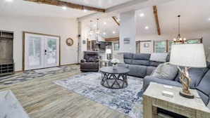Un salon spacieux avec un canapé gris, un fauteuil en cuir et une table basse ronde. Le sol est en bois et une grande fenêtre se trouve au fond de la pièce.