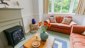 Una sala de estar acogedora con chimenea, un sofá, una mesa de centro con un jarrón y flores, y vistas a la vegetación a través del ventanal.