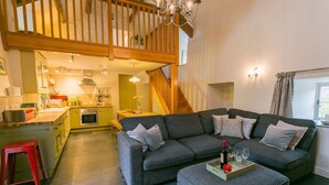 Un salon chaleureux avec un canapé gris, un escalier en bois et une cuisine en arrière-plan.