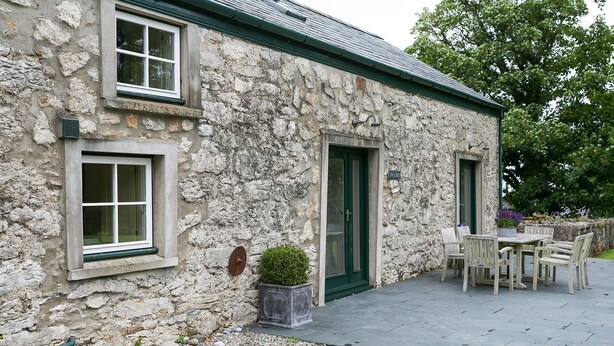 Un bâtiment en pierre avec deux fenêtres et une porte, un petit espace extérieur aménagé avec une table et des chaises, et un pot de fleurs.