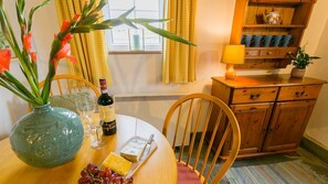 A dining table with a vase of flowers, a bottle of wine, glasses, cheese, and grapes. There is a wooden chair and a cabinet with a lamp.