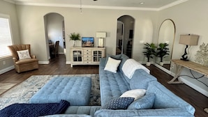Un vaste salon comprenant un grand canapé d’angle bleu, un fauteuil marron orné d’un coussin à motif de tigre, un meuble blanc aux portes en verre et un ventilateur de plafond.