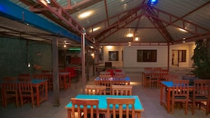 A covered dining area with wooden tables and chairs, a high ceiling with metal beams, and a stone wall on one side. - Taj Residency Pokhara (Pokhara)