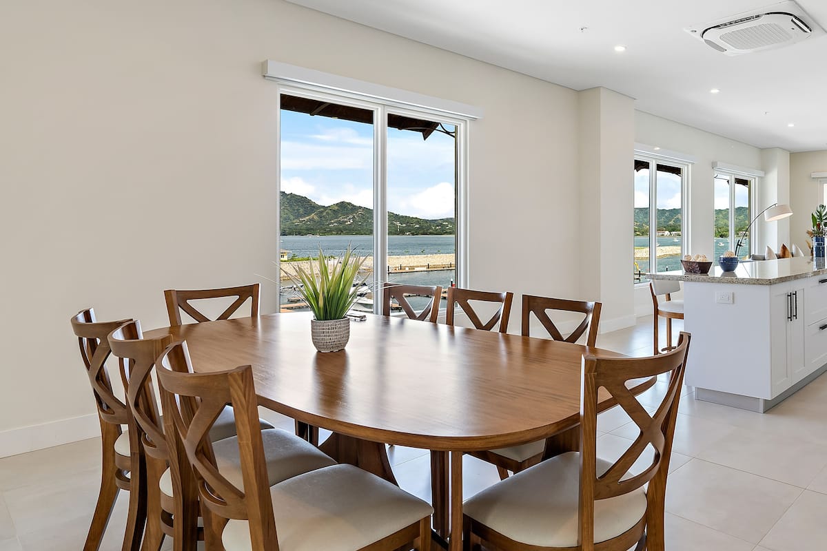 A dining area with a round wooden table and chairs, large windows with a view of a lake and hills, and a kitchen with white cabinets and a countertop.