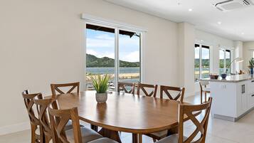Un comedor con una mesa redonda de madera y sillas, ventanas amplias con vista a un lago y colinas, y una cocina con armarios blancos y una encimera.
